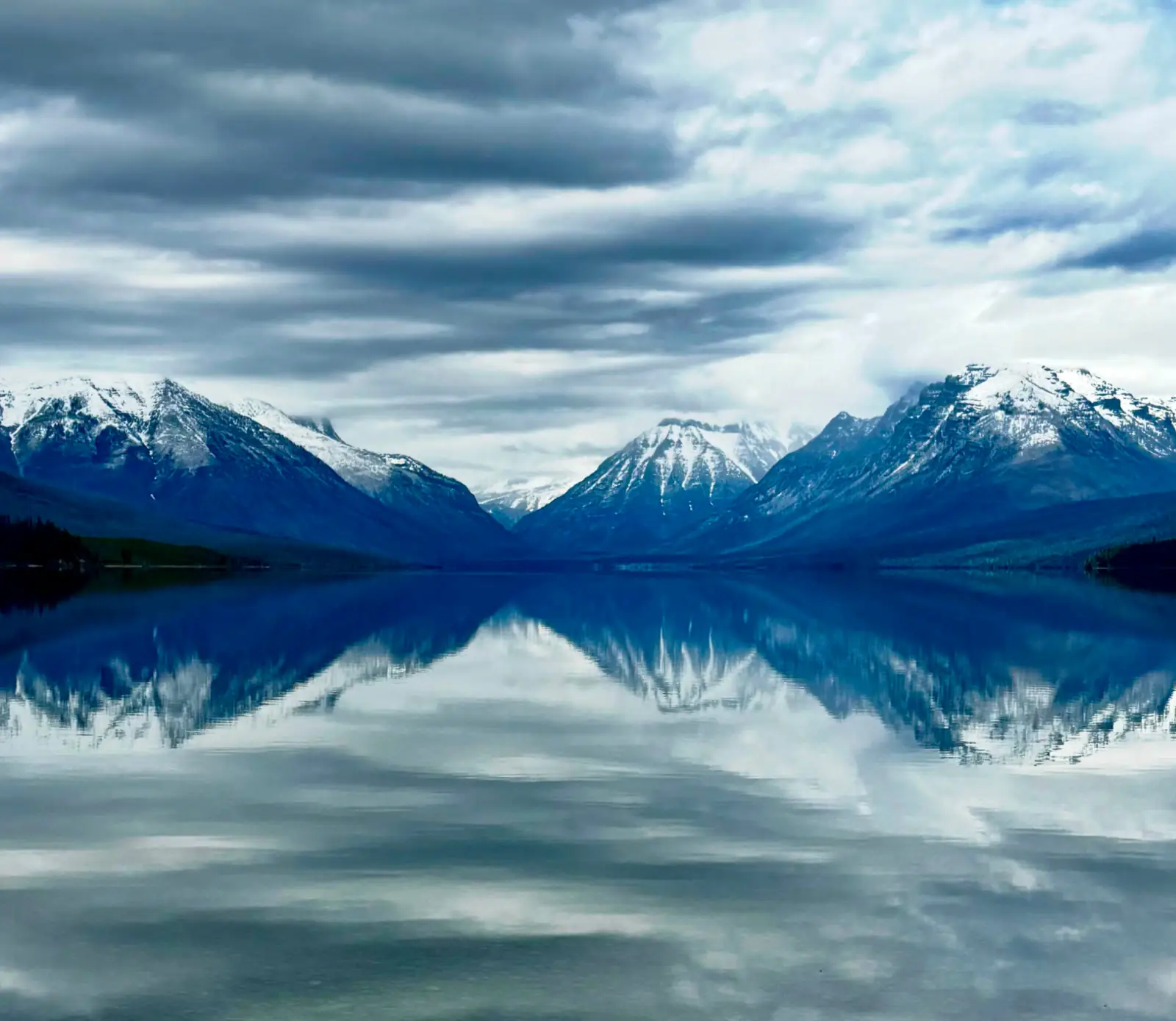 Snow-capped mountains mirrored in perfectly still water beneath a moody, cloud-streaked sky.