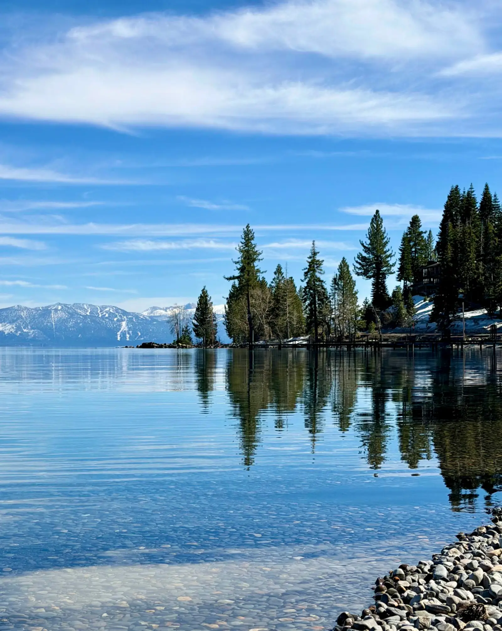 Pines on a small point reflected in calm blue water, snowy peaks in the distance.